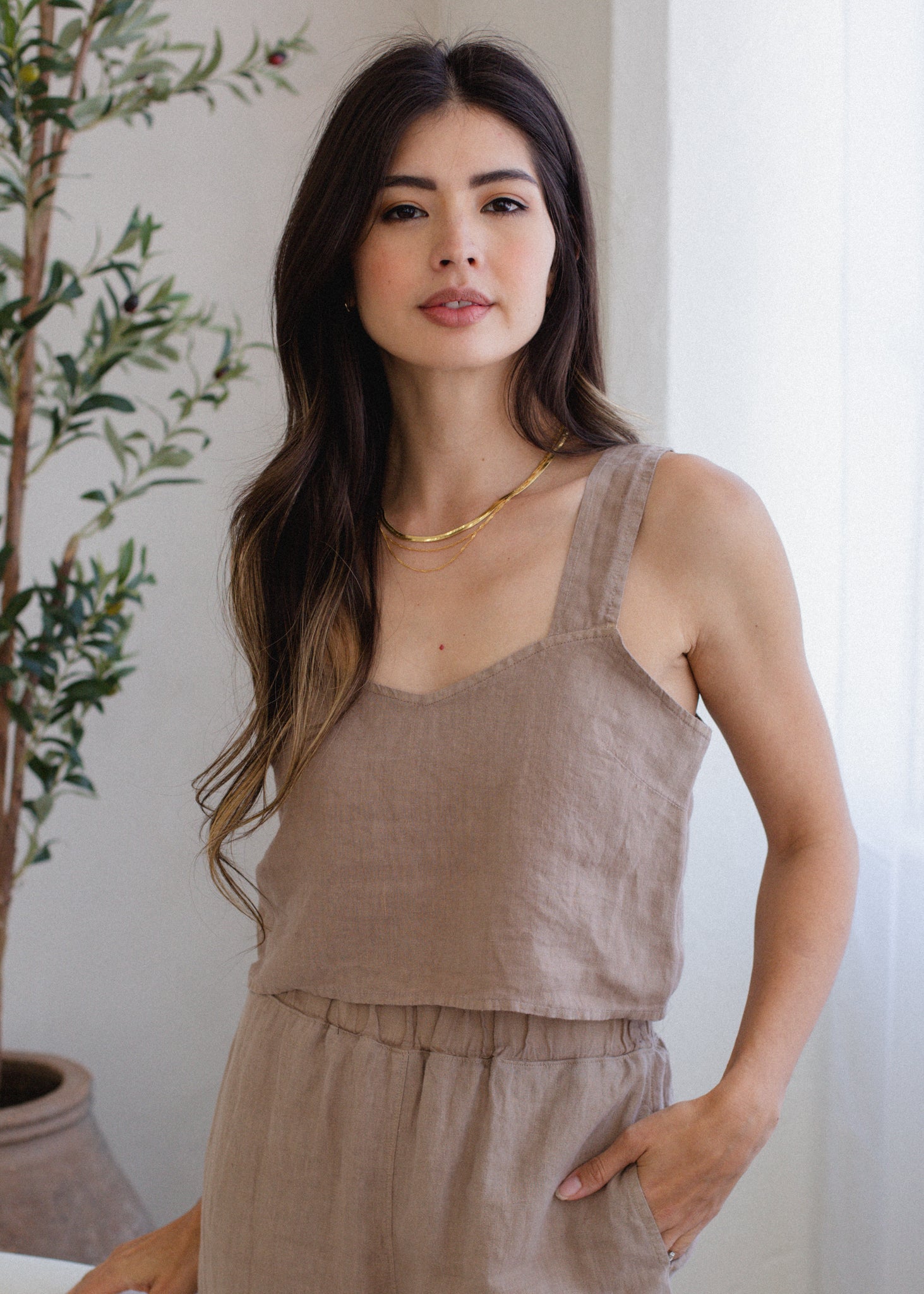 Woman wearing a beige sleeveless top and pants standing next to a plant indoors.