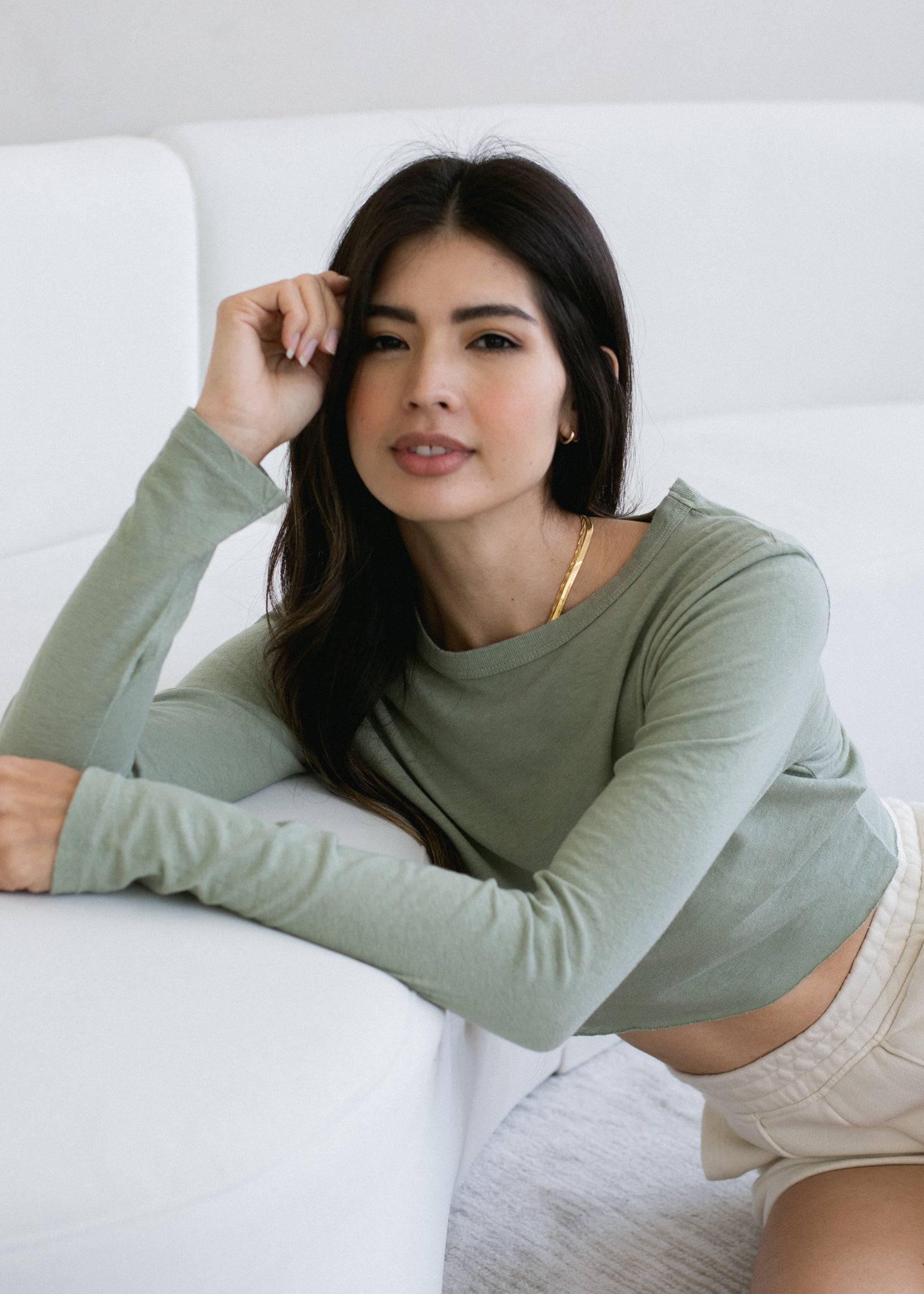 Woman wearing a green long-sleeve top sitting on a white couch.