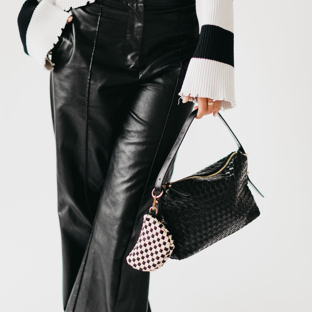 Person wearing black leather pants and holding a black handbag with a white and black checkered Mini Westlyn Bag Charm – Clip-On Mini Bag Accessory for Cash, Cards & AirPods