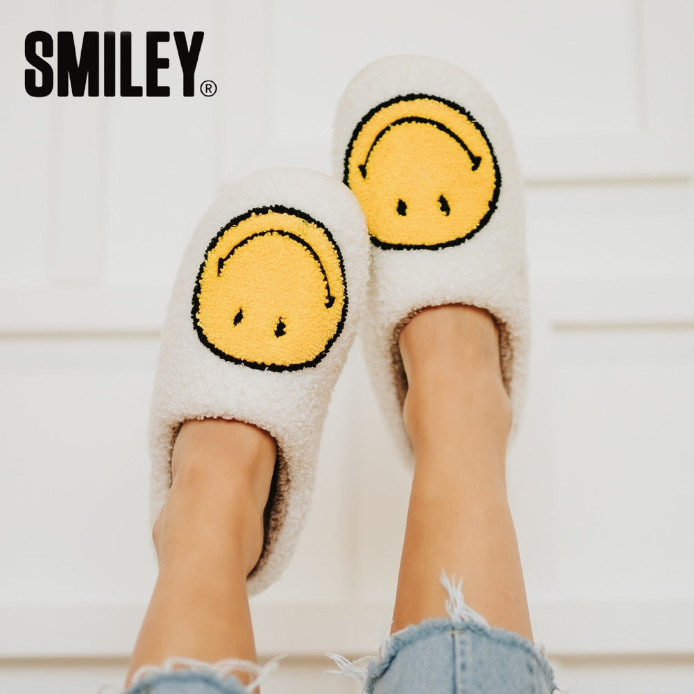 White fuzzy slippers with yellow smiley face design worn by a person on a light wooden floor.