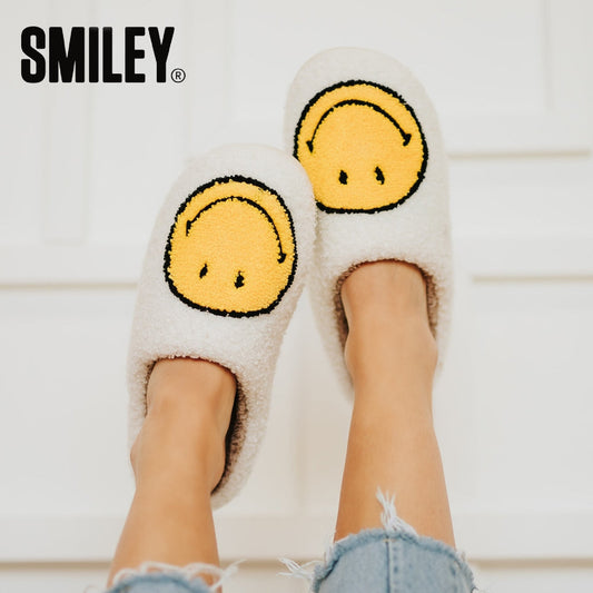 White fuzzy slippers with yellow smiley face design worn by a person on a light wooden floor.