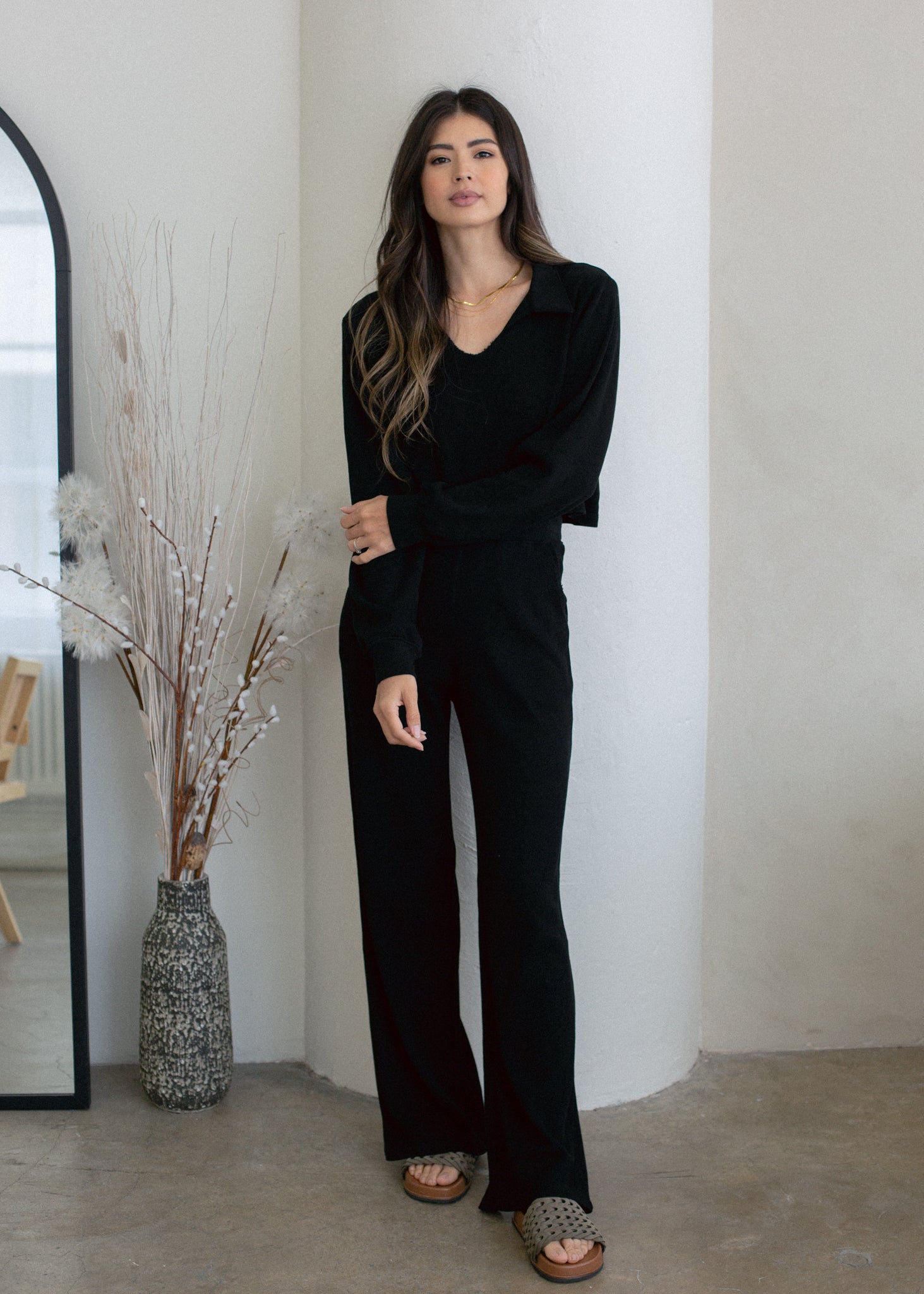 Woman wearing a black outfit standing in a minimalistic room with a vase and mirror.