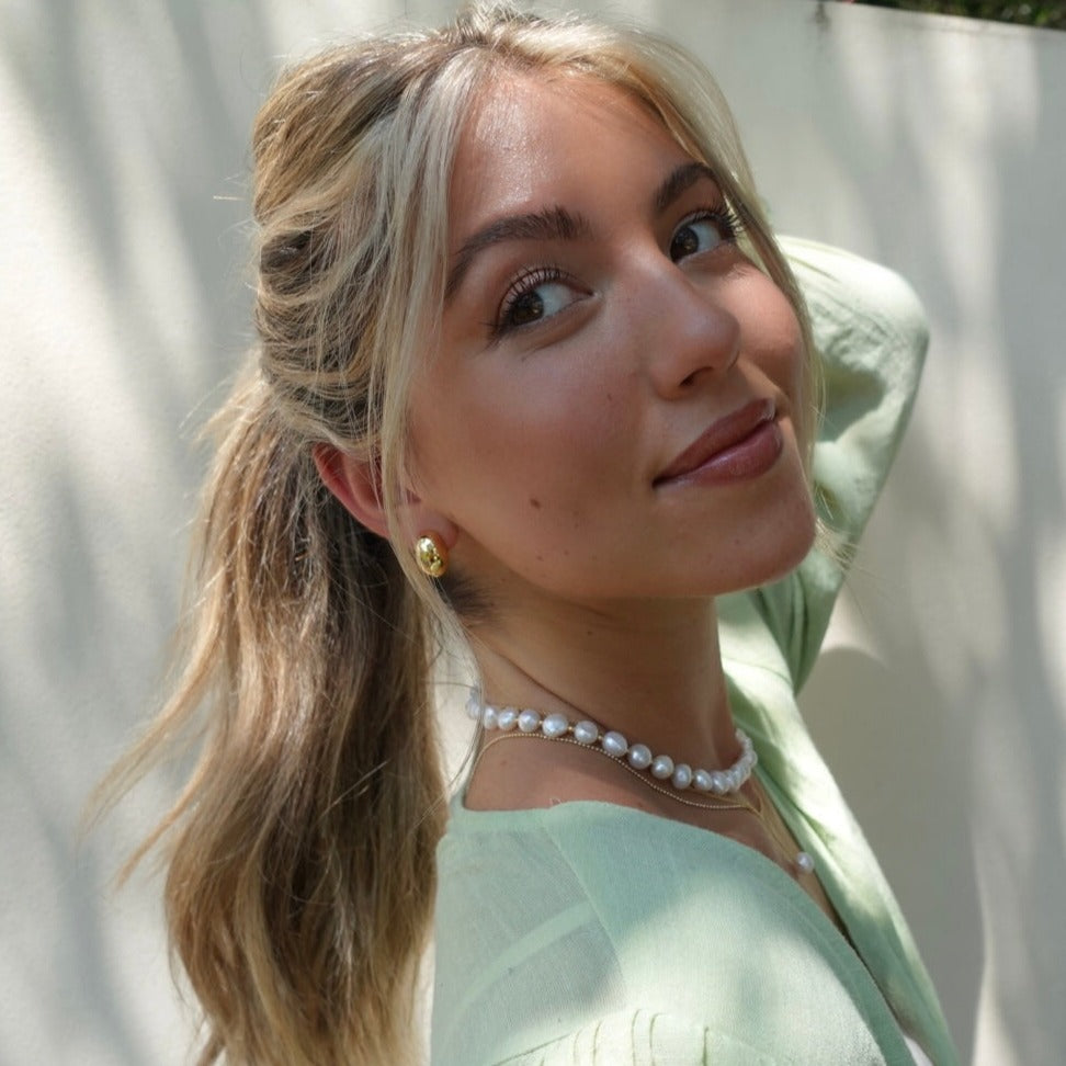 Woman with blonde hair tied back, wearing a pearl necklace and earrings, against a light background.