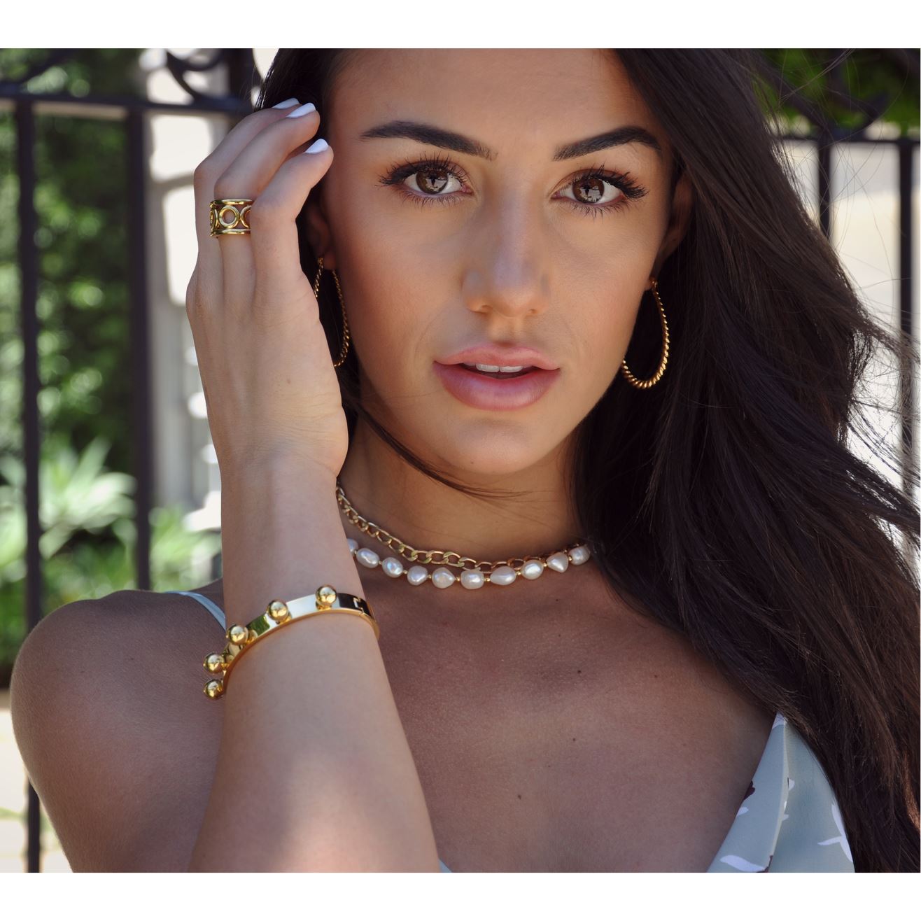 Woman wearing gold jewelry including a ring, bracelet, and necklace with a blurred outdoor background