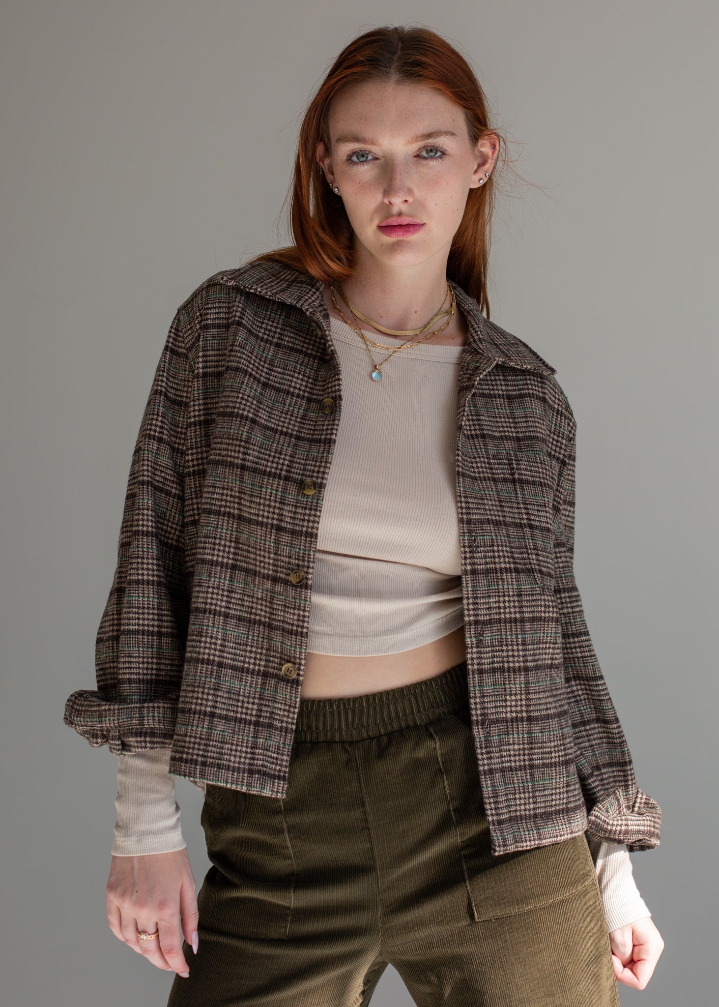 Woman wearing a plaid shirt over a beige top and green pants on a gray background