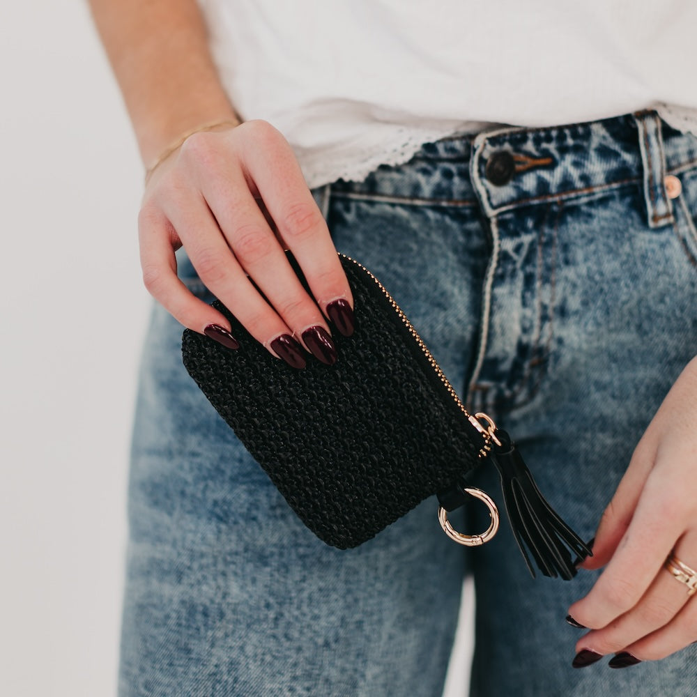Person holding a black woven clutch with a tassel detail against a neutral background