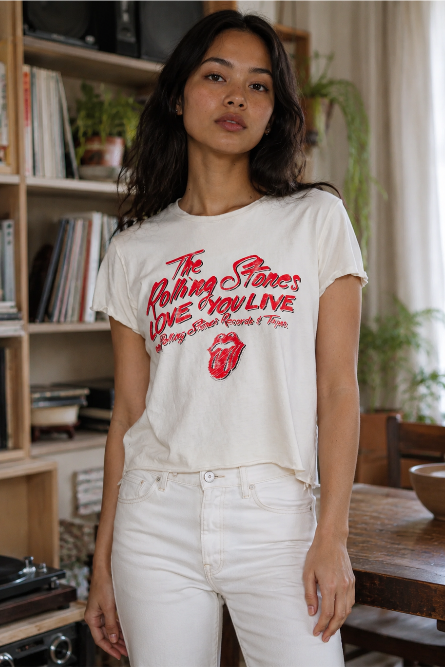 Woman wearing a Rolling Stones t-shirt in a room with a bookshelf and plants.