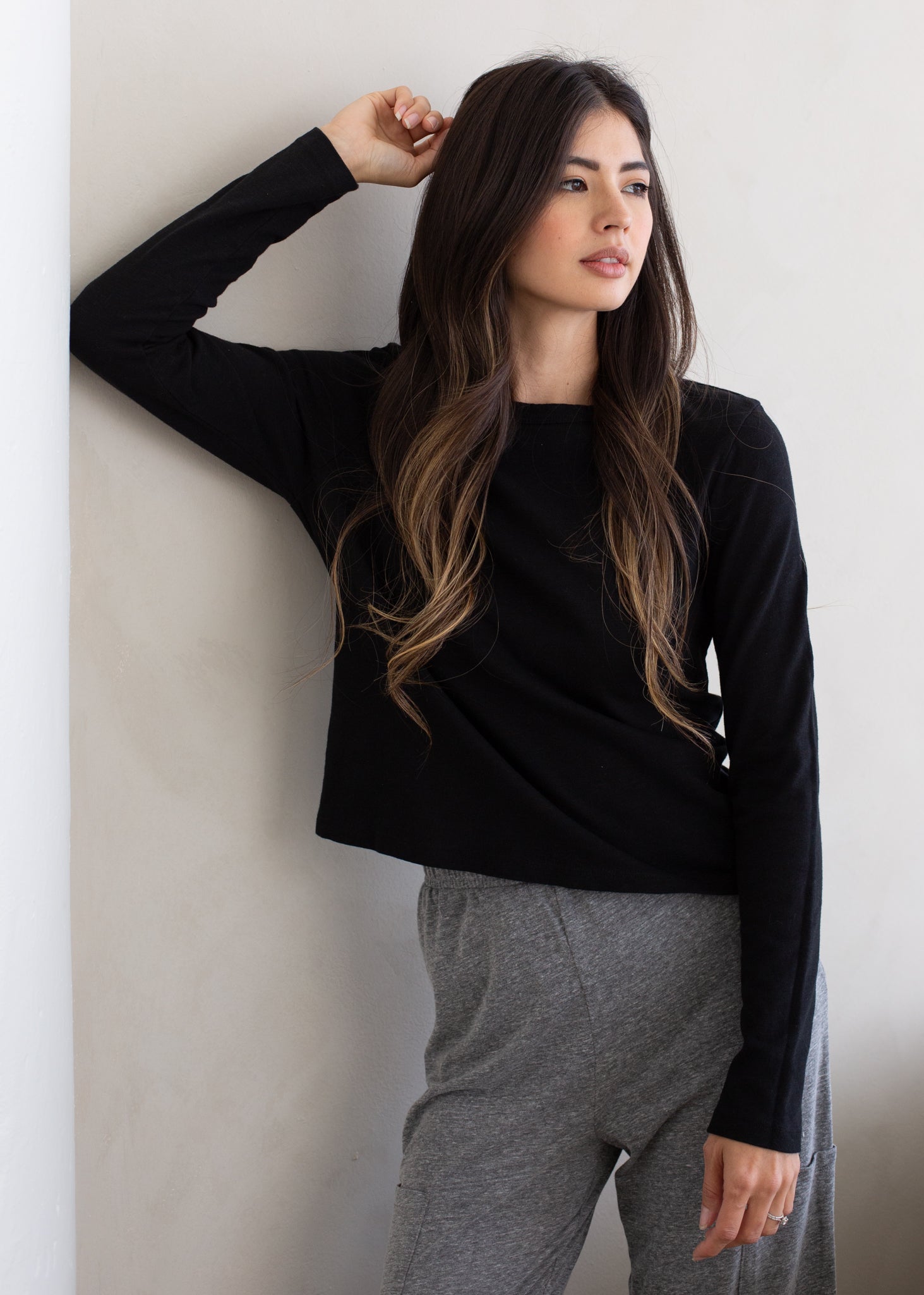 Woman wearing a black long-sleeve top and gray sweatpants against a white wall.