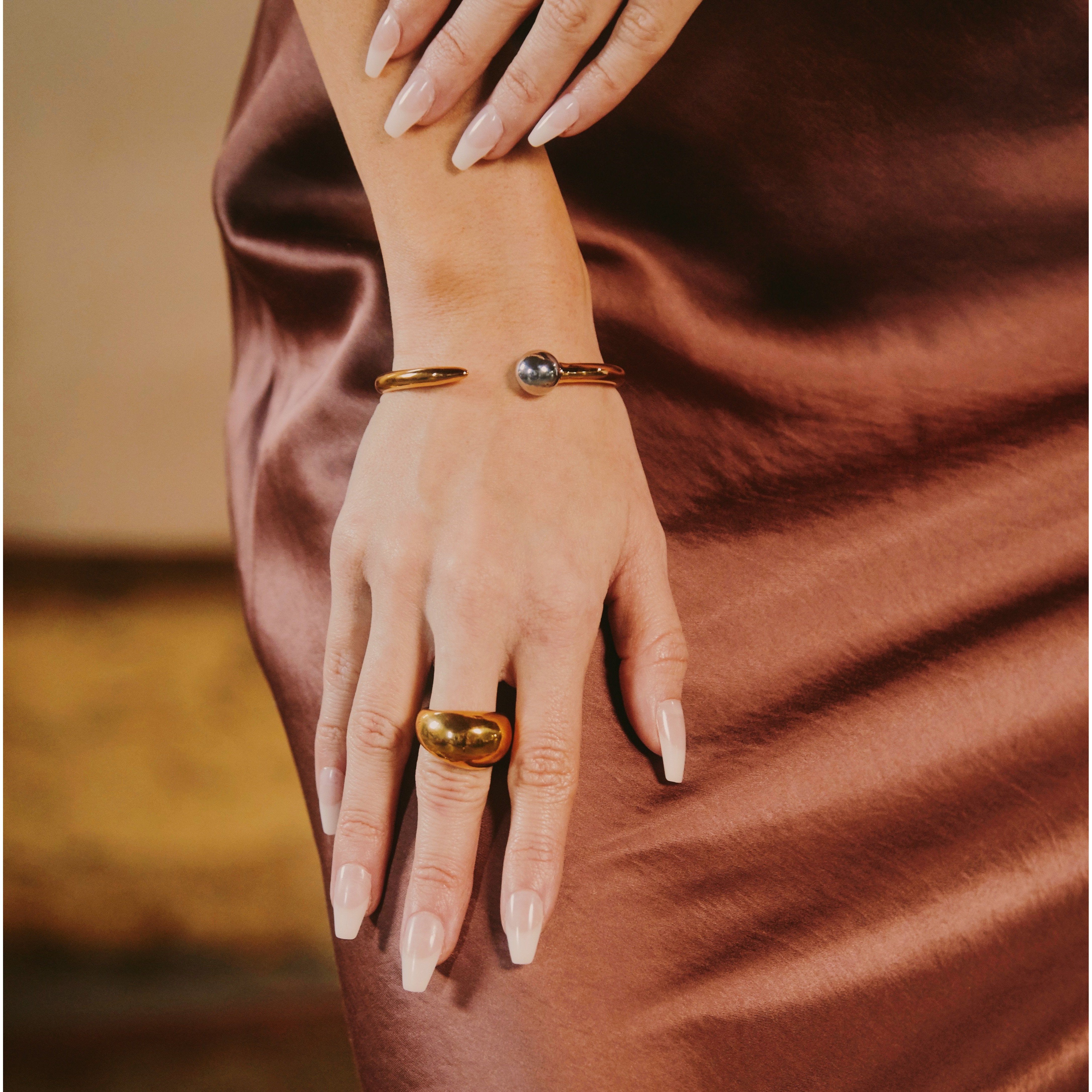Hand wearing gold braceler and ring on a brown fabric background