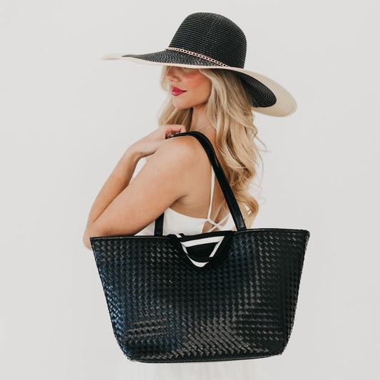 Woman wearing a black straw hat and holding a matching black straw tote bag on a light gray background