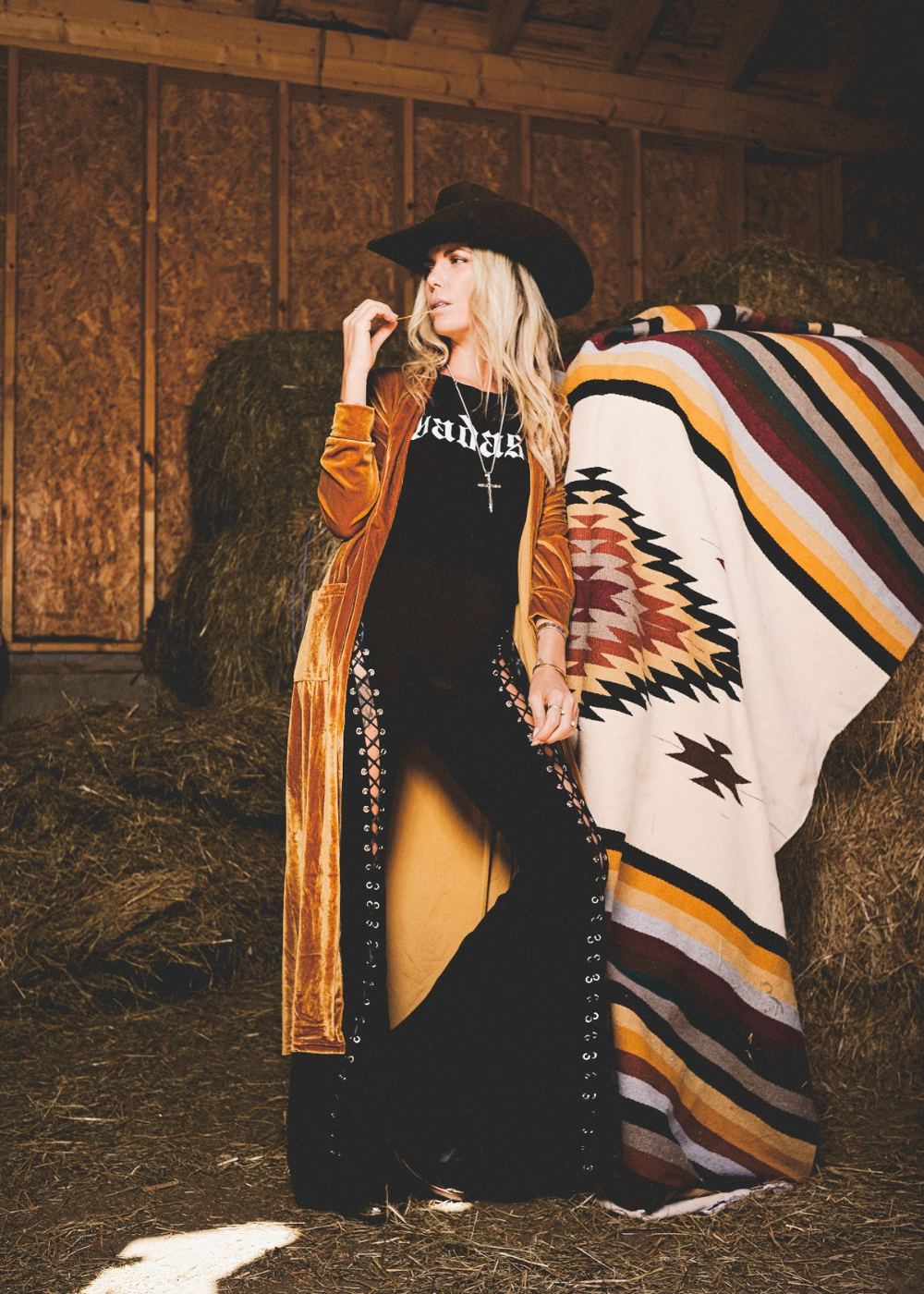 Woman in a cowboy hat and long coat standing next to a colorful patterned blanket in a barn setting.