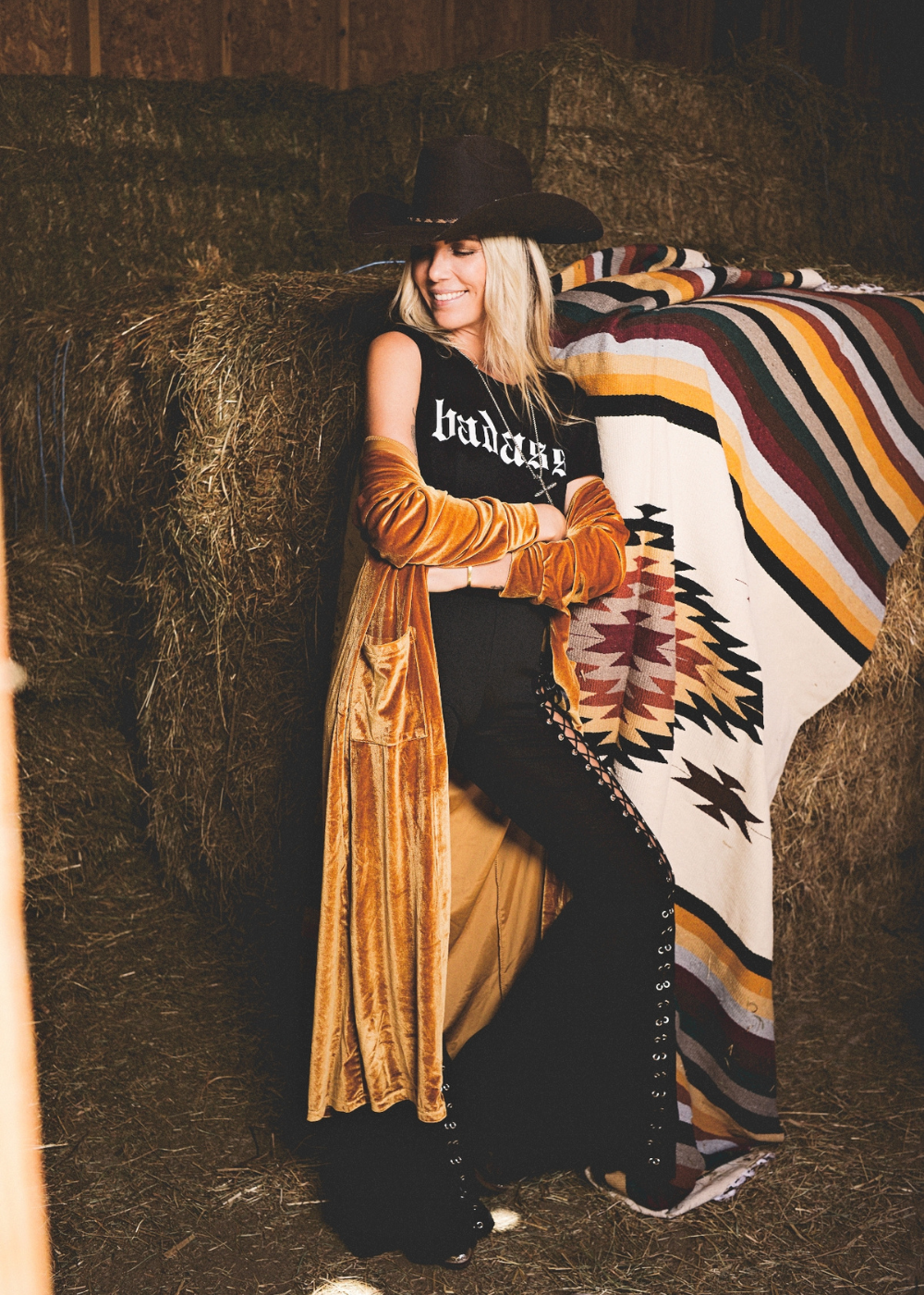Person in a black hat and long coat sitting on hay with a patterned blanket