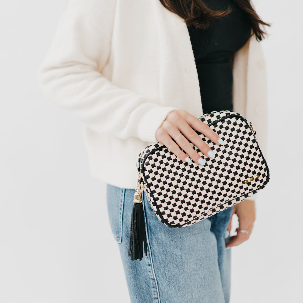 Person holding a black and white checkered clutch bag with a tassel on a plain background